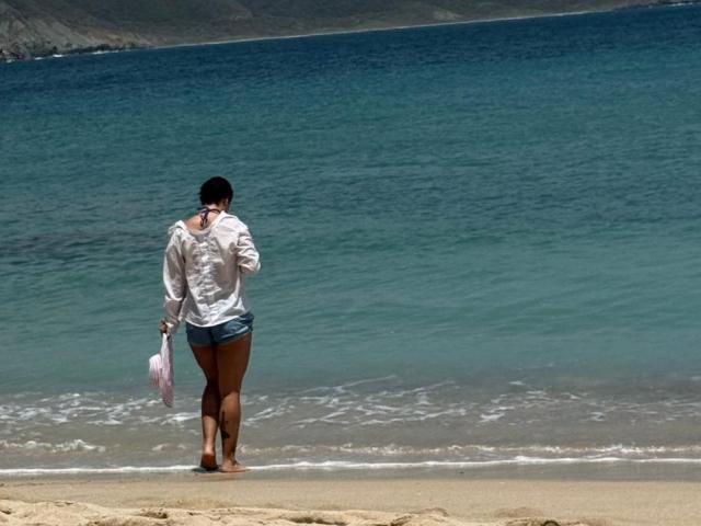 JuliaGarciax, le dos tourné vers la caméra, porte une chemise blanche et un short bleu, tient un sac rose et se tient debout sur une plage de sable avec de l'eau turquoise..