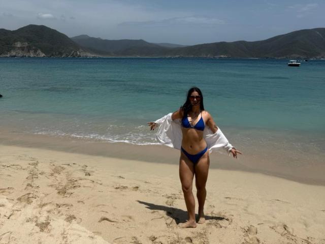 JuliaGarciax staat op een zandstrand, gekleed in een blauw bikini en een open wit shirt, met haar armen gespreid, waarbij haar borsten en billen zichtbaar zijn, met rode zonnebril, en erachter groen water..