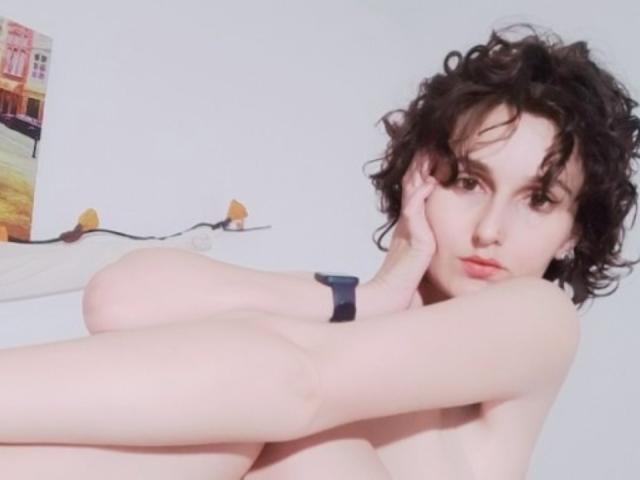 AnneRoss with curly black hair, pale skin, and a black wristband, is topless, covering her small tits with her hand, looking at the camera with a serious expression.