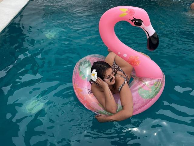 AlessiaFerry, con un seno di media grandezza, capelli scuri e un fiore bianco tra i capelli, è sdraiata su un gonfiabile a forma di fenicottero rosa in una piscina blu, indossando un bikini nero con stampa leopardo..