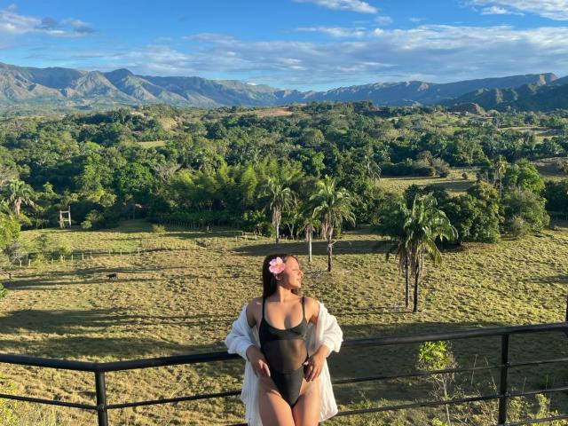 AlessiaFerry, con piel bronceada, traje de baño negro entero, bata blanca transparente, flor rosa en el cabello, de pie en el balcón, paisaje verde, palmeras, montañas, cielo azul claro, soleado, manos en las caderas..