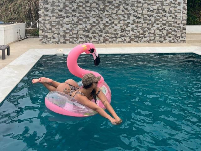 AlessiaFerry, butt up, in a pink flamingo float, blue-tinted pool, wearing a leopard print bikini and a leopard print hat, with her pussy partially visible.