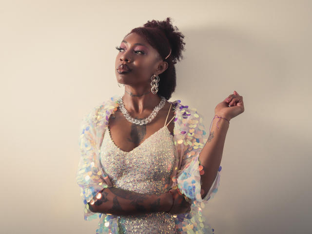 DaniaMarcell with dark skin, tattoos, pink eyeshadow, and a nose ring, wearing a sparkly dress and iridescent shawl, holding her hand up, looking to the side.