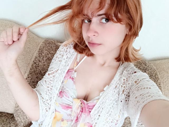 MireilleSpotwood met rood haar, die een haarstreng vasthoudt, draagt een witte kanten top over een roze bh met bloemenprint, waarbij haar kleine borsten zichtbaar zijn, lichte huid, een subtiele glimlach, zittend op een beige bank..