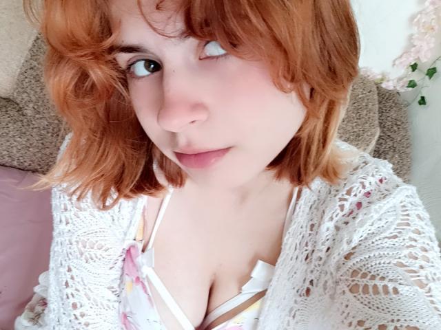 Mireille Spotwood, com cabelo ruivo, um cardigan de renda branca e decote visível com um sutiã floral, olhando para o lado com um leve sorriso. Fundo em tons suaves de rosa e bege..