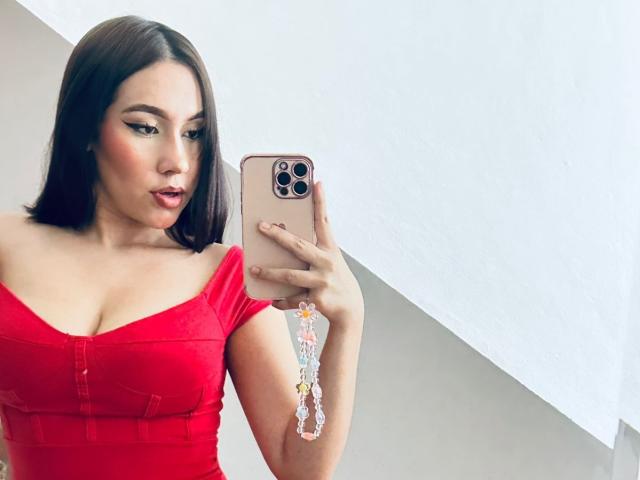 BibiSugarX with shoulder-length brown hair, red off-shoulder dress, holding a pink phone with a tassel case, showing her big tits and slightly parted red lips.