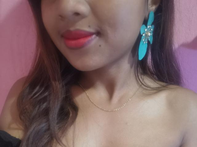 Shaniaha's face with red lipstick, blue flower earrings, and a gold necklace, showing a small mole on her upper lip, brown wavy hair, and a black off-shoulder top.
