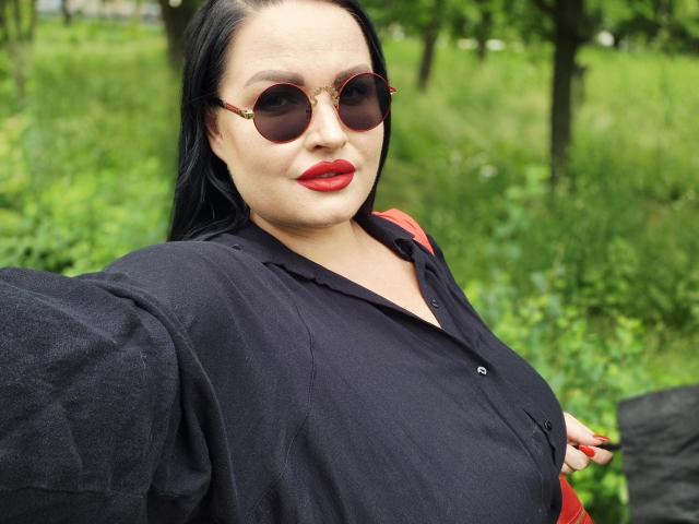 GeorgiaLerox mit schwarzem Hemd, rotem Lippenstift und goldfarbenen Sonnenbrillen, die eine rote Tasche hält, wobei ihre Brüste leicht sichtbar sind, dunkles Haar, grüner Rasen im Hintergrund, Selfie, selbstbewusst, sexy..