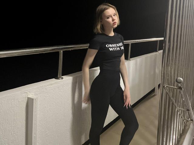 CheliaFloerke stands with a confident pose, wearing a black t-shirt and black pants, her ass slightly raised, and her tits small, in a dimly lit, metal-railled balcony.