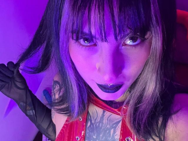 LiaMiss, con il viso illuminato di viola, rossetto nero e piercing al setto, indossa una giacca di pelle rossa, guanti neri e tiene i capelli. Tatuaggio visibile sul petto. Capelli tinti di nero e biondo..
