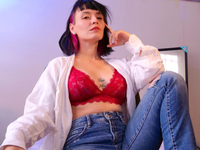SaraleePoison met zwart en roze haar, die een rode kantbeha, een wit shirt en blauwe jeans draagt, zit met één been opgetrokken, haar vinger op haar kin, en ziet er zelfverzekerd uit..