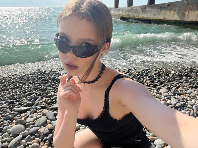 CheliaFloerke with brown hair, black sunglasses, black lace dress, and black choker, sitting on a rocky beach, hand near mouth, ocean and pier in background.