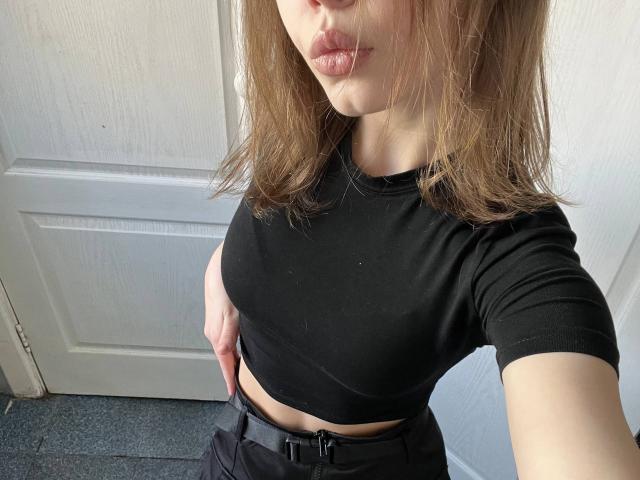 Ein Selfie von CheliaFloerke, auf dem sie ein schwarzes Crop-Top, kleine Brüste und schwarze Hosen mit einem Gürtel trägt. Ihr Gesicht zeigt geschmacklos hochgezogene Lippen, und sie hat braunes Haar. Im Hintergrund ist eine weiße Tür zu sehen..