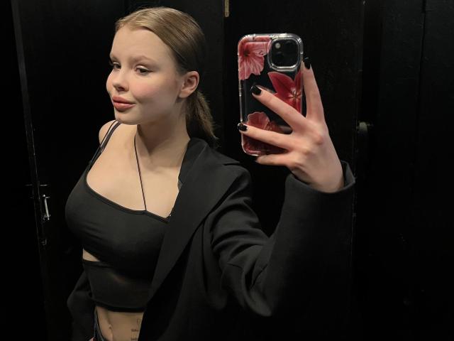 CheliaFloerke, con i capelli biondi raccolti in una coda di cavallo, indossa un top corto nero, una giacca nera e ha le unghie nere. Tiene in mano una cover per telefono con un motivo floreale rosso e sta scattando un selfie allo specchio, mostrando il seno..