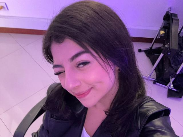 AnntonellaWatson has dark brown hair, a cute smile, and is wearing a black leather jacket. Her eyes are slightly winking, and she's in a tiled room with a camera.