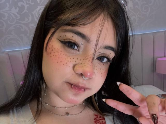 AmaraKazumi with freckles, nose ring, lip piercing, long dark hair, and winged eyeliner, making a peace sign, wearing a white top and multiple necklaces.