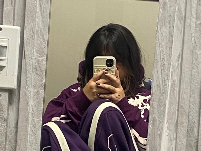 AmaraKazumi, con una sudadera y pantalones morados, sentada, tomando una selfie con el espejo usando un teléfono, cubriéndose la cara, mostrando cabello oscuro y ondulado, y esmalte de uñas morado..