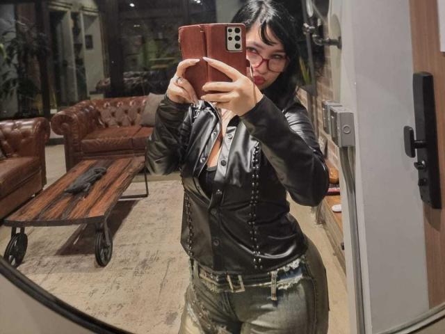 JulieTaylors with dark hair, red glasses, black leather vest, and ripped jeans, taking a mirror selfie in a dimly lit room. Boobs partially visible under the vest.