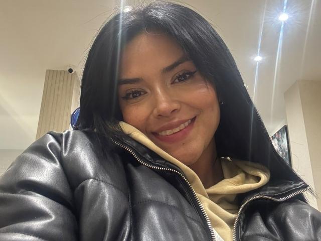 MajoJones, met een glimlach en zwart haar, draagt een zwarte leren jas over een beige hoodie. Ze heeft een schattig gezicht met bruine ogen en een kleine neus..