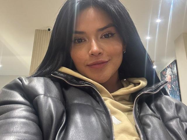 MajoJones, com cabelos longos e pretos, pele morena e maquiagem discreta, vestindo uma jaqueta preta acolchoada sobre um moletom bege, sorrindo suavemente, olhos castanhos e pequenos brincos de argola, em um quarto bem iluminado..