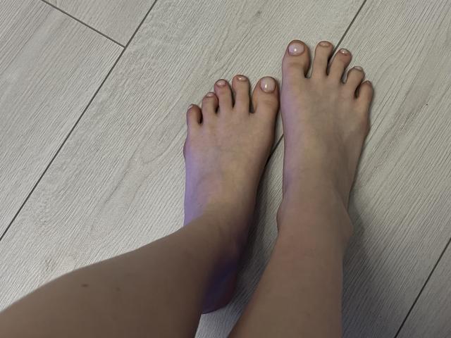 Ginnia's bare feet with light skin, slightly pinkish toes, and clean nails, standing on a light wooden floor. Her toes are slightly apart, showing a relaxed position.