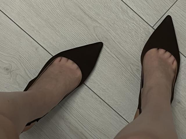 I piedi di Ginnia, calzati con tacchi a punta neri, mostrano leggermente le dita dei piedi incurvate, mentre è in piedi su un pavimento di legno bianco. Le sue gambe sono pallide e indossa collant trasparenti..