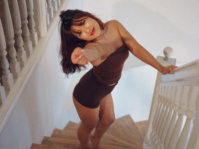 ErikaKim with red hair, brown strapless dress, and tan skin, pointing downward, standing on wooden stairs, hands on white rail, smiling, showing small tits and ass.