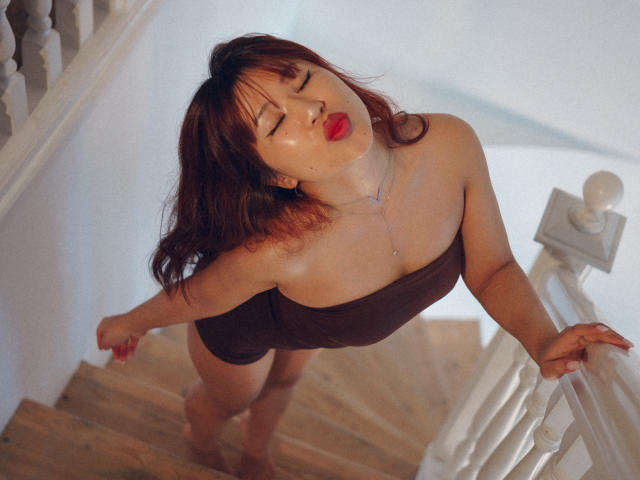 ErikaKim with red lipstick, eyes closed, leaning on a staircase, wearing a brown strapless top, showing her tits, and a necklace, with her ass slightly raised.