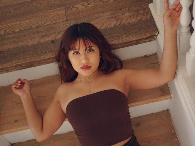 ErikaKim with red hair, brown strapless top, lying on wooden stairs, arm on white railing, looking up, red lipstick, necklace, wooden texture, relaxed, confident, sexy.
