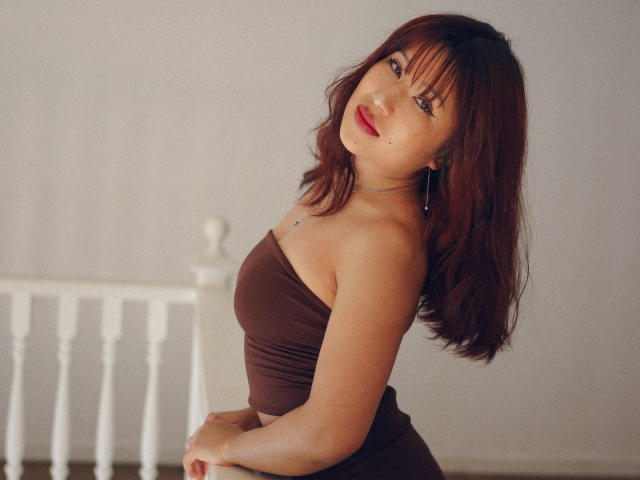 ErikaKim has red hair, wears a brown strapless top, and is looking back with a slight smile. She's holding her butt, showing her tits. Her tits are small.