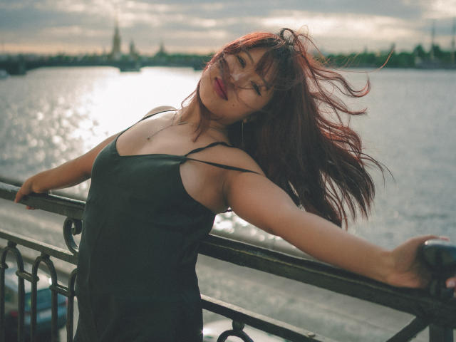 ErikaKim with red hair, black spaghetti strap dress, leaning on railing, sunlight in eyes, wind in hair, hands on railing, water and skyline in background.