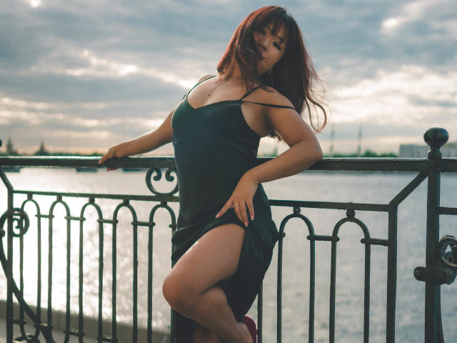 ErikaKim with red hair, wearing a black dress, standing on a balcony, raising her dress to show her thick, sexy thighs, and her small, perky tits, under a cloudy sky.