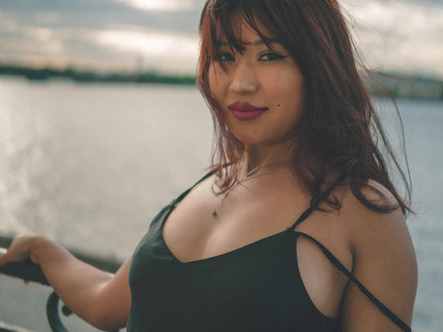 ErikaKim with red hair, dark green spaghetti strap top, showing her tits, standing by a waterway, smiling, pink lipstick, necklace, holding a railing, dusk sky in background.