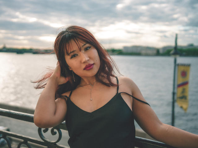ErikaKim with red hair, black spaghetti strap top, pink lipstick, and a small nose piercing, standing by a waterfront, hand in hair, water reflecting in the background.