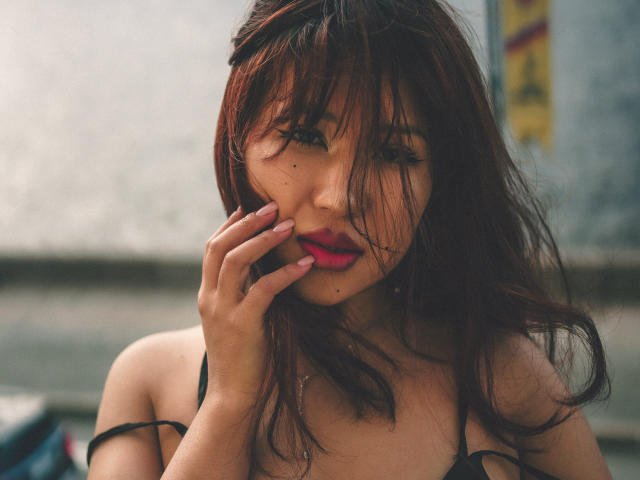 ErikaKim with dark, wet hair, red lipstick, and black strap dress, touching her slightly parted, pink-lipped, mocha-skinned face, showing small beauty mark on her right cheek.