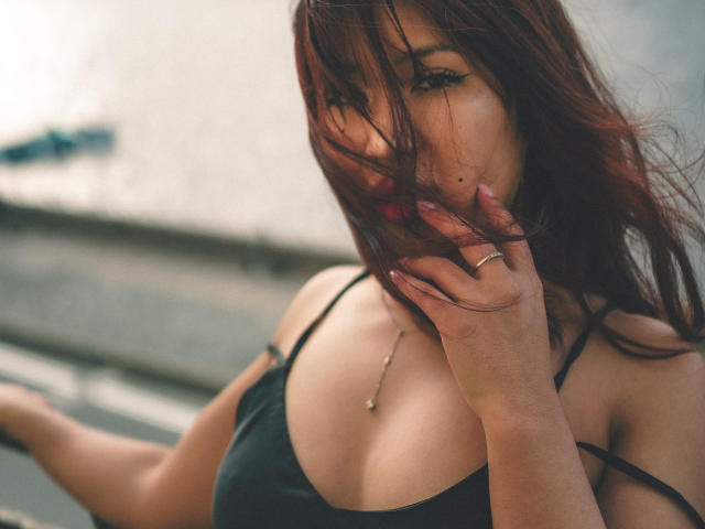 ErikaKim with red hair, black spaghetti strap top, hand covering her mouth, showing her tits, small silver necklace, and ring, looking seductively at the camera.
