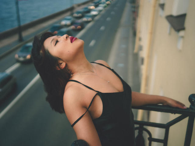 ErikaKim with her head tilted back, eyes closed, wearing a black spaghetti-strap dress, red lipstick, and a small necklace, leaning on a railing, surrounded by a street with cars.