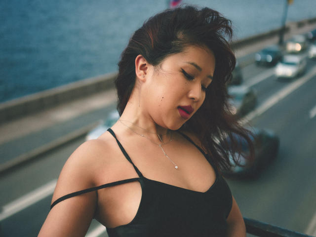 ErikaKim with dark hair, wearing a black dress with side cutouts, showing her tits, necklace, and red lipstick, standing by a street with blurred cars in the background.