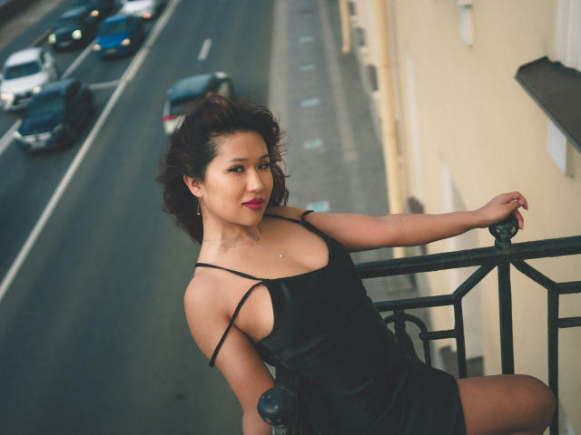 ErikaKim with dark curly hair, black strap dress, leaning on balcony, showing off her tits, red lipstick, and a slight smile, cars in background, urban setting.