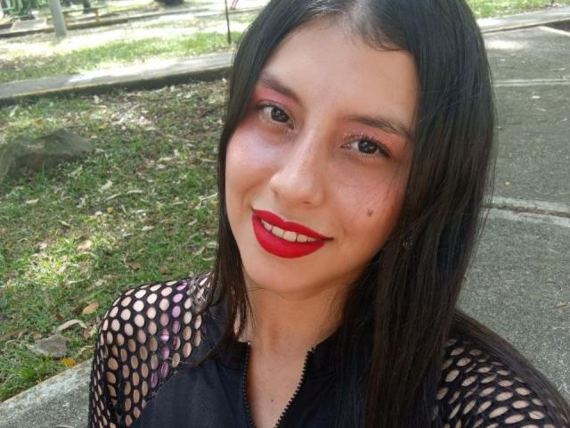 MadissonFontaine with red lipstick, black fishnet top, and long brown hair, smiling at the camera, showing her shiny brown eyes and small beauty mark on her left cheek.