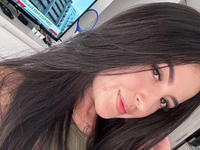 SofiaLopezHot has long black hair, brown eyes, and a small red blemish on her cheek. She's wearing a green top and a gold necklace, smiling softly.