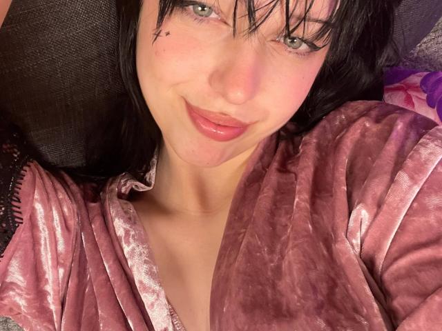 AmaiRin, com o cabelo preto molhado, um robe de veludo rosa e um sorriso discreto, exibindo seus seios e lábios rosados, está deitada em uma superfície escura. Sua pequena marca na região púbica é visível..