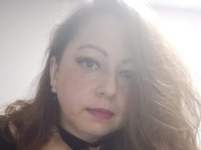 CristelleDeLuxe has light skin, brown wavy hair, and pink lipstick, wearing a black choker and earrings, with a slightly blushed face and subtle makeup.