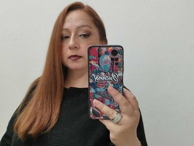 CristelleDeLuxe with red hair, black sweater, holding phone with colorful case, wearing dark lipstick, ring on finger, looking at mirror, showing phone to camera.