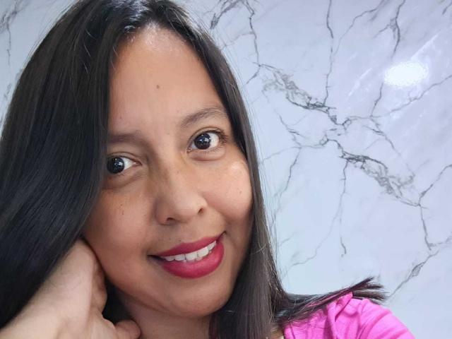 AngelJazmin, com cabelo preto e liso, tom de pele médio, batom vermelho, sorrindo, tocando sua bochecha, vestindo uma camisa rosa, parede de mármore ao fundo..