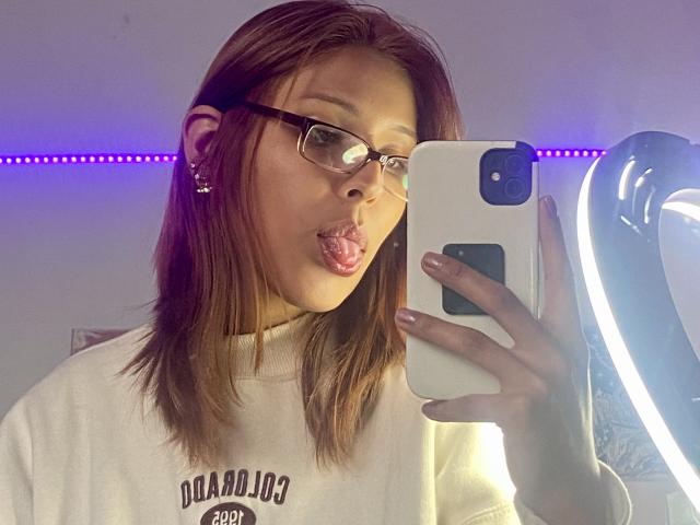 KiaraHernandezX with brown hair, glasses, and a white hoodie, pouting her lips, holding a white iPhone, standing in front of a ring light with purple LED strip.