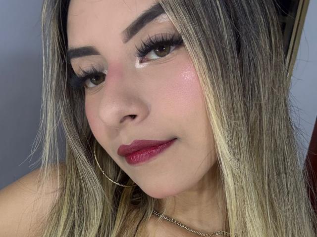 CloeRouss has blonde hair, thick eyebrows, long lashes, pink blush, shiny skin, red lipstick, and a subtle smile, wearing a thin necklace and gold hoop earrings.