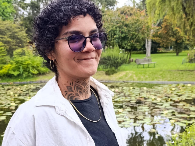 AnaRoa, con i capelli ricci e tatuaggi sul collo, indossa occhiali da sole, una camicia bianca e una t-shirt nera, e sorride alla fotocamera. Ha il seno piccolo e un viso carino..