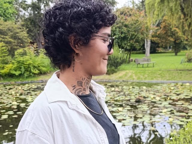 AnaRoa, com cabelo preto encaracolado, óculos, brincos de argola, tatuagem no pescoço, camisa branca, camiseta preta, está em pé perto de um lago de lírios. Seus seios são pequenos, e ela está sorrindo..