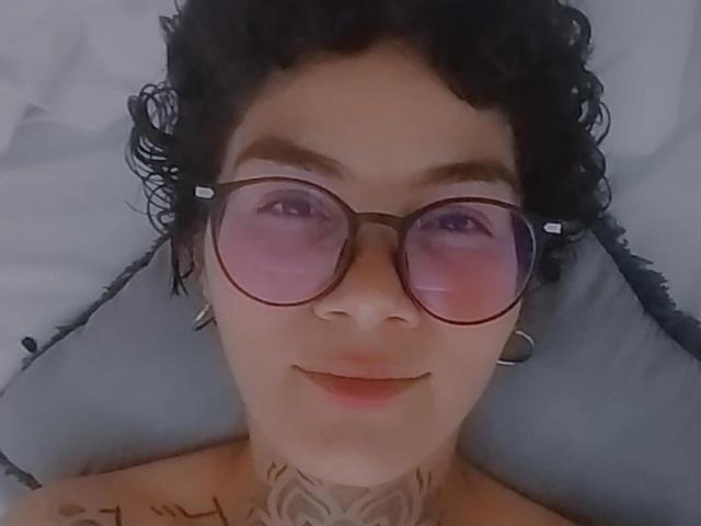 AnaRoa, con i capelli neri ricci, che indossa occhiali con una leggera sfumatura rosa e piccoli orecchini a cerchio, sdraiata, mostra tatuaggi sul collo e sul petto, sorridendo dolcemente..